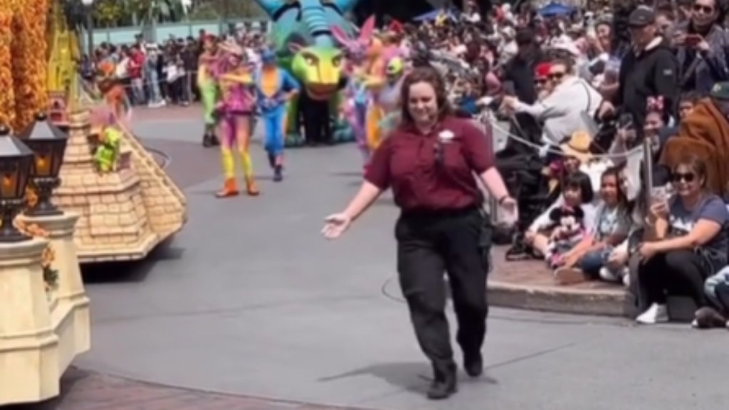 A Disney park parade is paused. The crowd looks on at a Disney employee who is walking up to something that can't be seen in this image, arms open wide