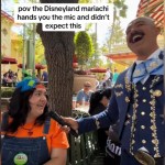 A woman sits at a table. A man in a mariachi band stands near her, holding a mic up to her. She sings and the man looks amazed, mouth agape