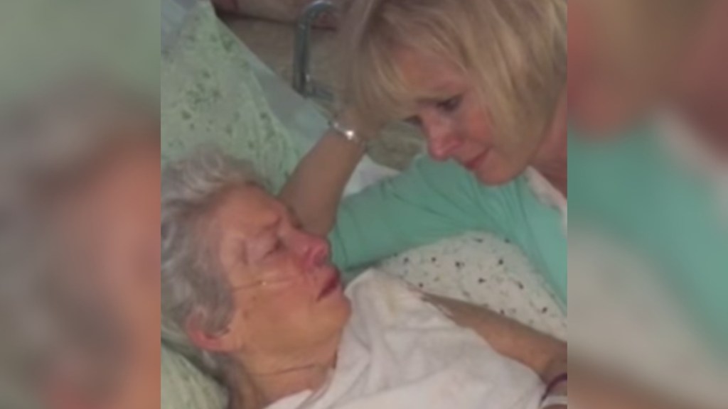 An elderly woman lays in bed. Her adult daughter lays next to her, propped up on her shoulder. The woman looks down at her mom as she speaks