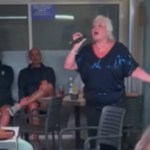 A woman stands at the center of a room, various people sitting at tables around her. She holds a mic in one hand as she stretches out her other arm wide. She sings passionately into the mic. People nearby look amazed