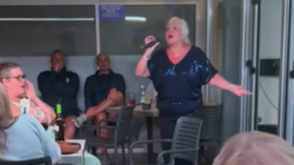 A woman stands at the center of a room, various people sitting at tables around her. She holds a mic in one hand as she stretches out her other arm wide. She sings passionately into the mic. People nearby look amazed