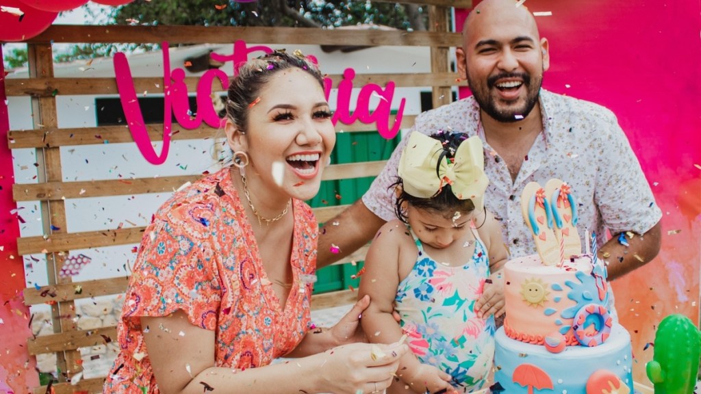 A man and woman stand outside with their little girl. They stand behind a table with a colorful beach-themed birthday cake. They adults are smiling wide and the girl looks down at her bathing suit