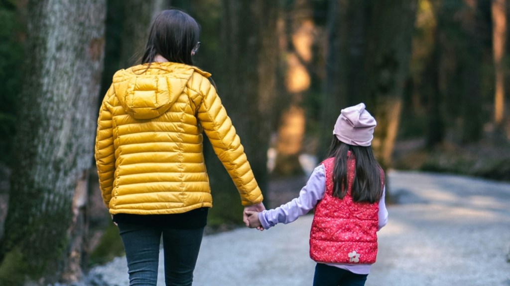 View from behind of a woman and her little girl holding hands as they take a walk outside on a path in nature