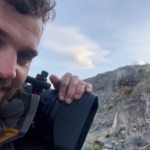 Close up of a man holding a professional camera with his one free hand. Just a few feet behind him is a puma looking off in the distance