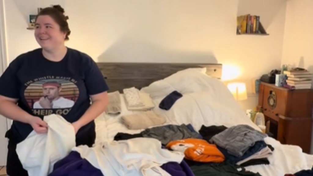 A woman stands next to a bed full of laundry. She holds a shirt in her hand, but she's looking off to the side. She's smiling and her mouth agape because she's impressed.