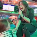 A singing waitress leans down from her small stage to hold hands with a little girl sitting at a table. The woman sings into a mic. Other patrons look over at them both, smiling