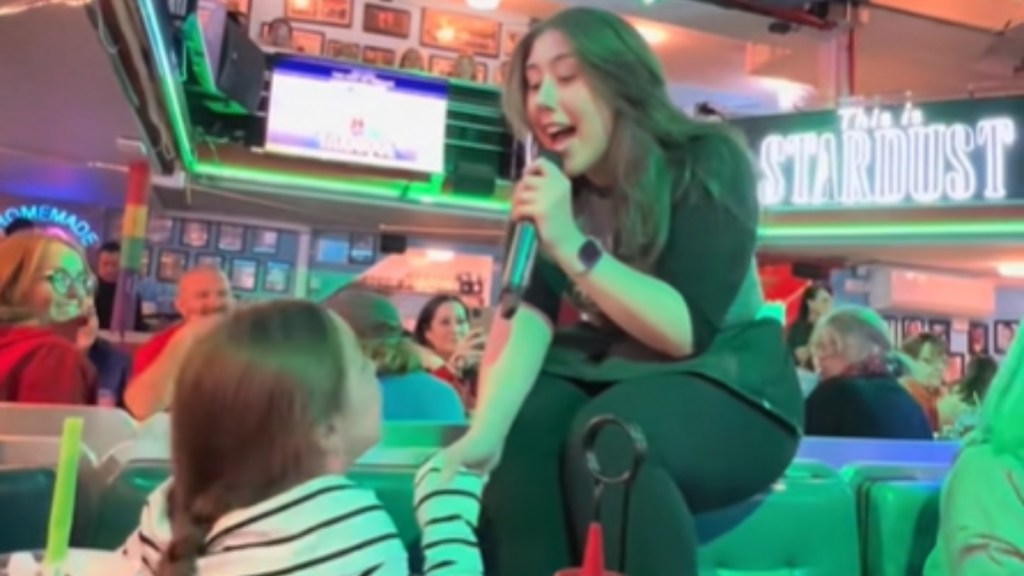A singing waitress leans down from her small stage to hold hands with a little girl sitting at a table. The woman sings into a mic. Other patrons look over at them both, smiling