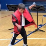 A little boy dressed like Michael Jackson in the "Thriller" music video dances. He holds a mic in his raised hand.