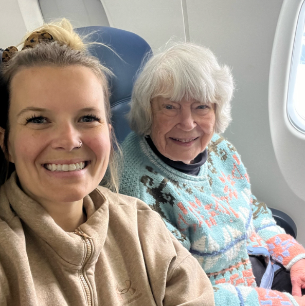 A woman, Riley, smiles as she takes a photo in a plane. Next to her is an elderly woman, Mary, who smiles wide.