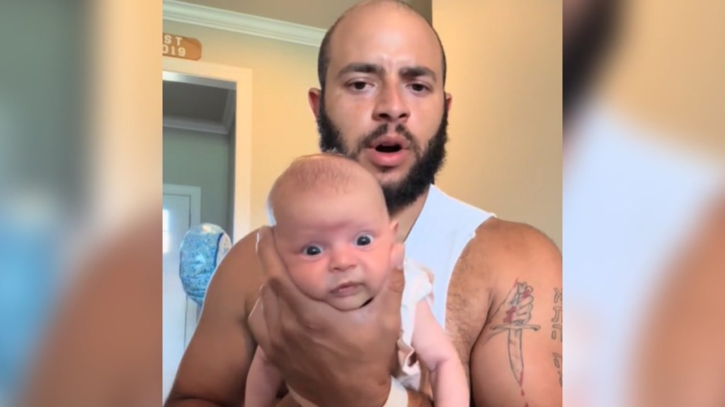 A man talks as he holds his newborn in his hands. The baby's head rests between his thumb and pointer finger. The baby looks wide-eyed at the camera